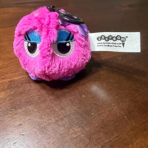 May include: A pink plush toy with large blue eyes, a purple bow, and a black clip. The toy has a fluffy texture and a tag that reads "iscream". The toy is on a wooden surface.
