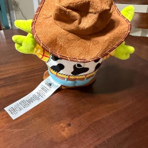 May include: A plush toy of a cowboy character, featuring a brown hat, yellow shirt, and a cow-print vest. The toy has green arms and a blue belt. A white tag with text is attached.