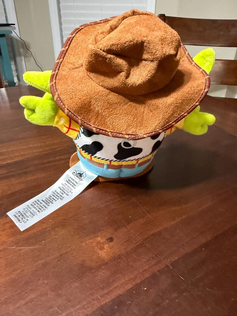May include: A plush toy of a cowboy character, featuring a brown hat, yellow shirt, and a cow-print vest. The toy has green arms and a blue belt. A white tag with text is attached.