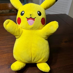 May include: A plush Pikachu toy in a standing pose with arms outstretched. The toy is primarily yellow with black-tipped ears, red cheek circles, and a pink mouth. The toy is made of soft fabric.