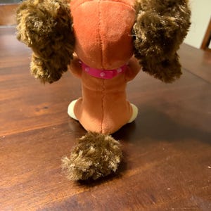 Pode incluir: Um c&atilde;o de pel&uacute;cia, visto por tr&aacute;s, com um corpo castanho, orelhas grandes e fofas e uma cauda a condizer. O c&atilde;o usa uma coleira rosa com bolinhas brancas. O brinquedo est&aacute; sobre uma superf&iacute;cie de madeira escura.