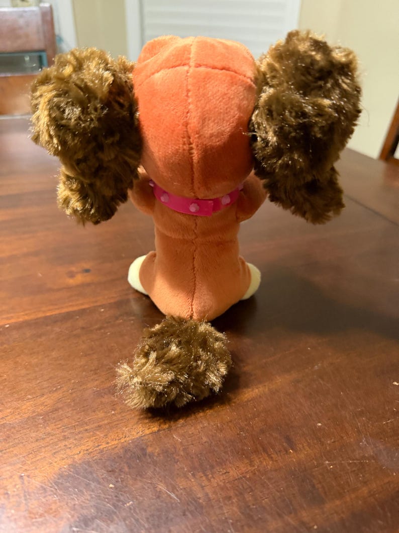 Pode incluir: Um c&atilde;o de pel&uacute;cia, visto por tr&aacute;s, com um corpo castanho, orelhas grandes e fofas e uma cauda a condizer. O c&atilde;o usa uma coleira rosa com bolinhas brancas. O brinquedo est&aacute; sobre uma superf&iacute;cie de madeira escura.