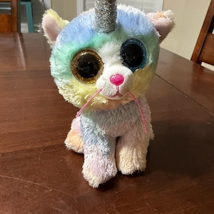 May include: A plush unicorn cat toy with a rainbow-colored head and body. It has large, glittery eyes, a pink nose, and a silver horn. The toy is sitting on a wooden surface.