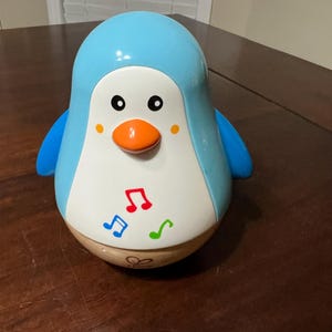 May include: A blue and white penguin-shaped toy with an orange beak and black eyes. The toy has blue wings and musical notes on its belly. The base of the toy is a light brown color.