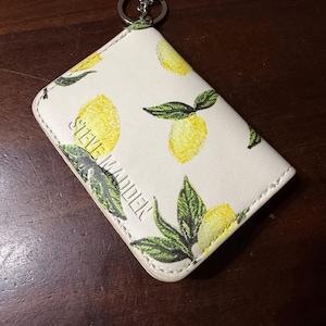 May include: Cream-colored cardholder with a lemon pattern and the text "STEVE MADDEN". The cardholder has a silver key ring attached. The lemons are yellow with green leaves.