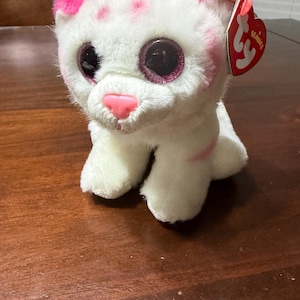 Puede incluir: Un gato de peluche blanco con detalles rosas en las orejas, la cara y el cuerpo. El gato tiene ojos grandes y brillantes y una nariz rosa. Una etiqueta roja en forma de corazón está unida a la oreja.