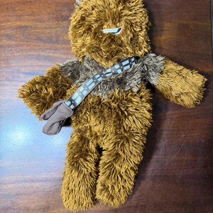 May include: A plush toy depicting a character with brown and tan fur, a bandolier, and a small pouch. The toy has a detailed face with a white eye and a gray nose. The arms and legs are also detailed.