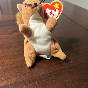 May include: A brown plush squirrel toy with a white belly and a red heart-shaped tag. The tag has a yellow star and the word "Ty" printed on it. The squirrel has black eyes and whiskers.