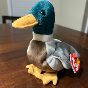 May include: A plush duck toy with a teal head, yellow beak and feet, and a brown and gray body. The toy has a white collar and a red and white tag attached to its side. The duck is sitting on a wooden surface.