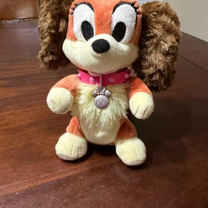Pode incluir: Um brinquedo de pel&uacute;cia de cachorro com pelo laranja e creme, orelhas marrons e olhos pretos grandes. Ele usa uma coleira rosa com uma etiqueta em forma de pata que diz "Fifi". O brinquedo est&aacute; em uma superf&iacute;cie de madeira.