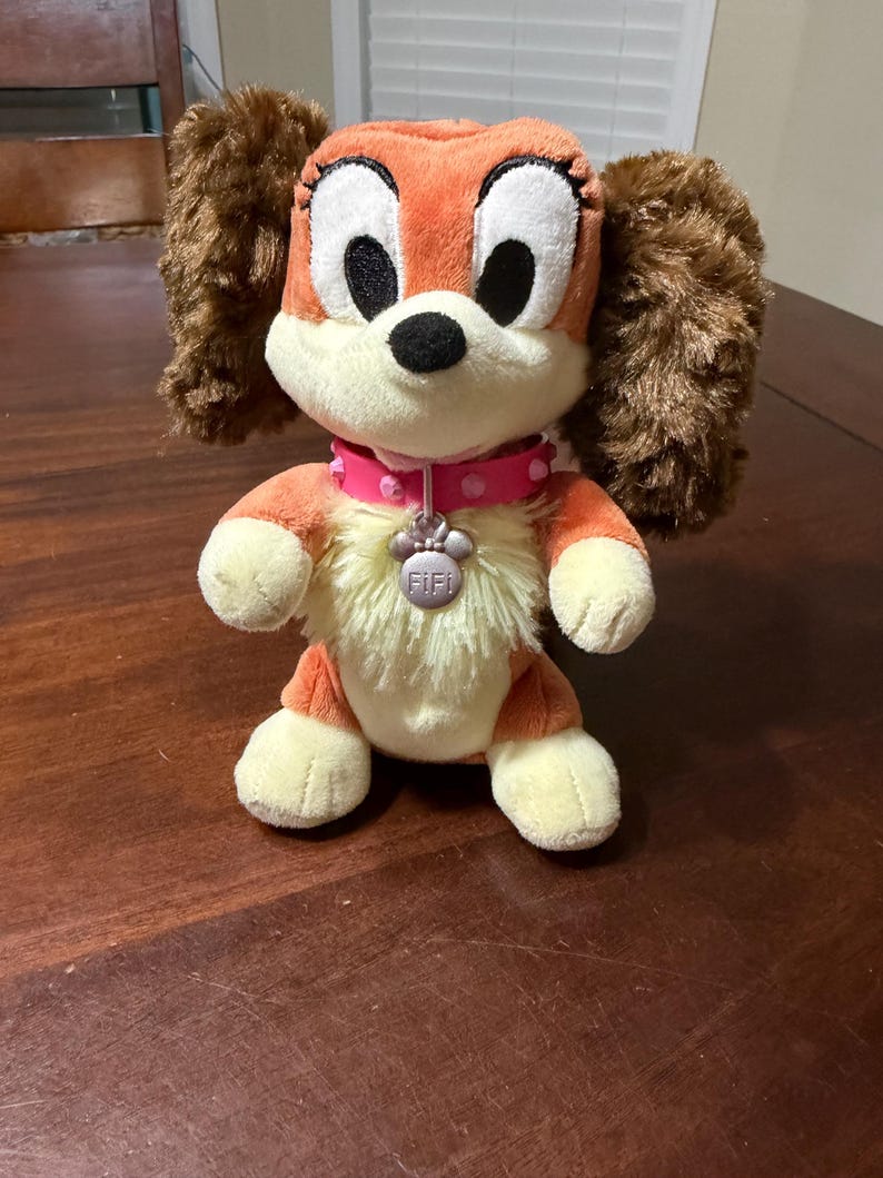 Pode incluir: Um brinquedo de pel&uacute;cia de cachorro com pelo laranja e creme, orelhas marrons e olhos pretos grandes. Ele usa uma coleira rosa com uma etiqueta em forma de pata que diz "Fifi". O brinquedo est&aacute; em uma superf&iacute;cie de madeira.
