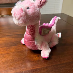 May include: A plush pink dragon toy with a sparkly, textured neck and accents. The dragon has a soft, fluffy body, small wings, and a friendly expression. The ears and wings have a glittery pink pattern.