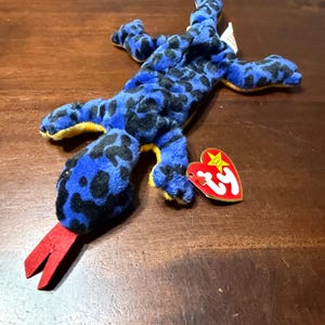 May include: A blue plush lizard toy with black spots and yellow accents. The lizard has a red tongue and a heart-shaped tag attached. The toy is lying on a wooden surface.