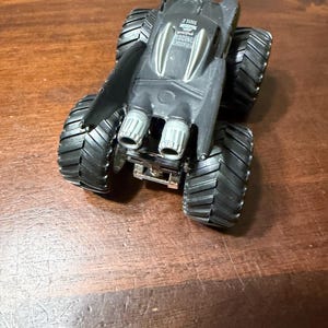 Puede incluir: Un cami&oacute;n monstruo de juguete negro con neum&aacute;ticos grandes y texturizados. El cami&oacute;n tiene un dise&ntilde;o elegante con tubos de escape plateados y el texto "2 TIME RACING CHAMPION". El juguete es de pl&aacute;stico y parece ser un art&iacute;culo de colecci&oacute;n.