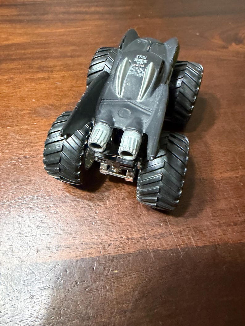 Puede incluir: Un cami&oacute;n monstruo de juguete negro con neum&aacute;ticos grandes y texturizados. El cami&oacute;n tiene un dise&ntilde;o elegante con tubos de escape plateados y el texto "2 TIME RACING CHAMPION". El juguete es de pl&aacute;stico y parece ser un art&iacute;culo de colecci&oacute;n.