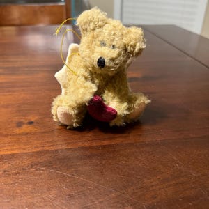May include: A small, plush teddy bear ornament with a light brown fur texture. The bear has a black nose and is holding a small, red heart. A gold string is attached for hanging, and a white star-shaped element is behind the bear.