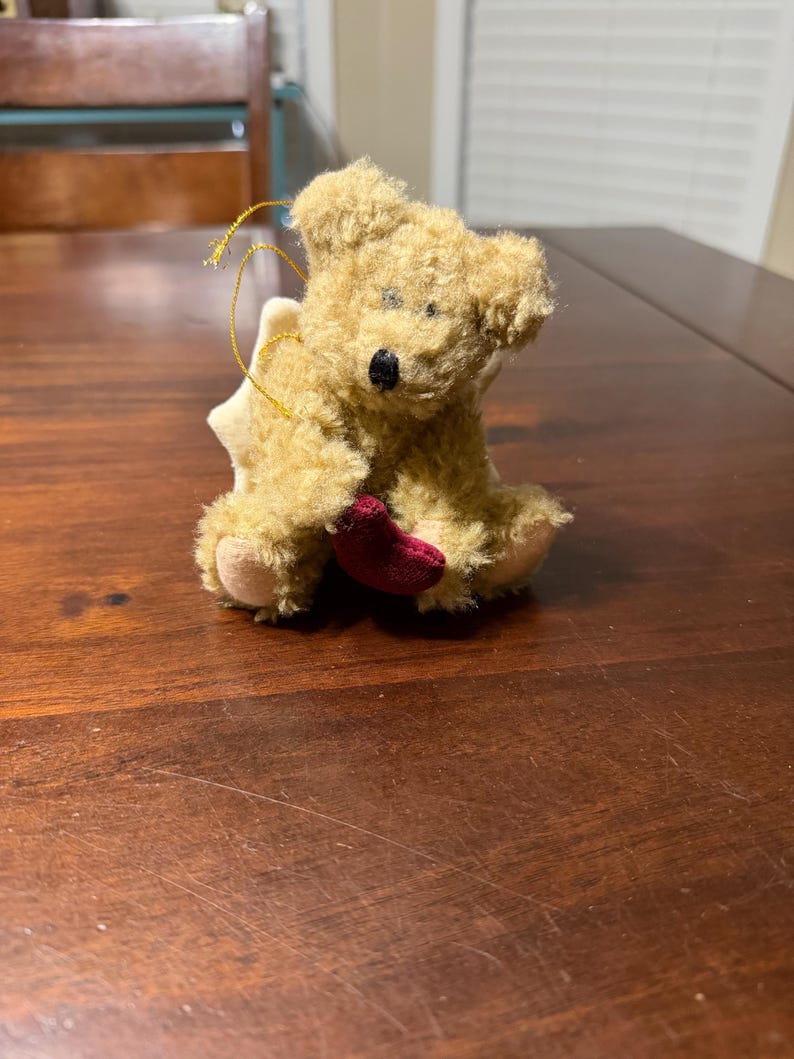 May include: A small, plush teddy bear ornament with a light brown fur texture. The bear has a black nose and is holding a small, red heart. A gold string is attached for hanging, and a white star-shaped element is behind the bear.