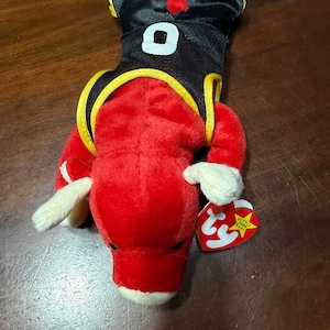 May include: A red plush toy shaped like a bull, wearing a black jersey with the number 0. The jersey has yellow trim. The toy has white hooves and a red tag.