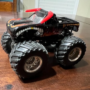 Op de afbeelding: Zwart monstertruck speelgoed met grote, getextureerde banden en een gedetailleerde carrosserie. De truck heeft een dreigend gezichtsontwerp met een grijnzende mond en rode accenten. De tekst "El Toro Loco" is zichtbaar.