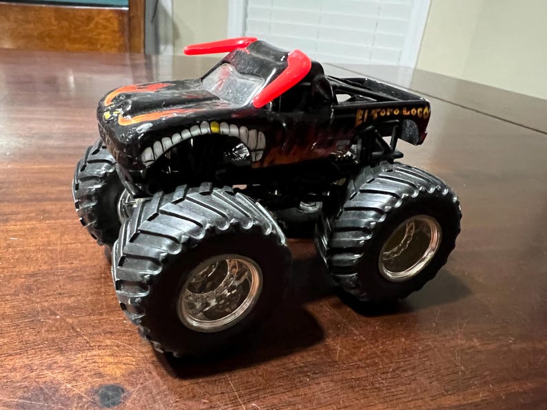 Op de afbeelding: Zwart monstertruck speelgoed met grote, getextureerde banden en een gedetailleerde carrosserie. De truck heeft een dreigend gezichtsontwerp met een grijnzende mond en rode accenten. De tekst "El Toro Loco" is zichtbaar.