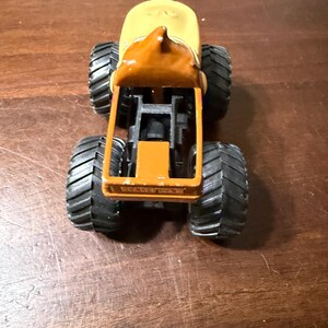 May include: A toy monster truck with a yellow cab and a brown roof. The truck has large black tyres and a brown chassis. The truck is sitting on a wooden surface.