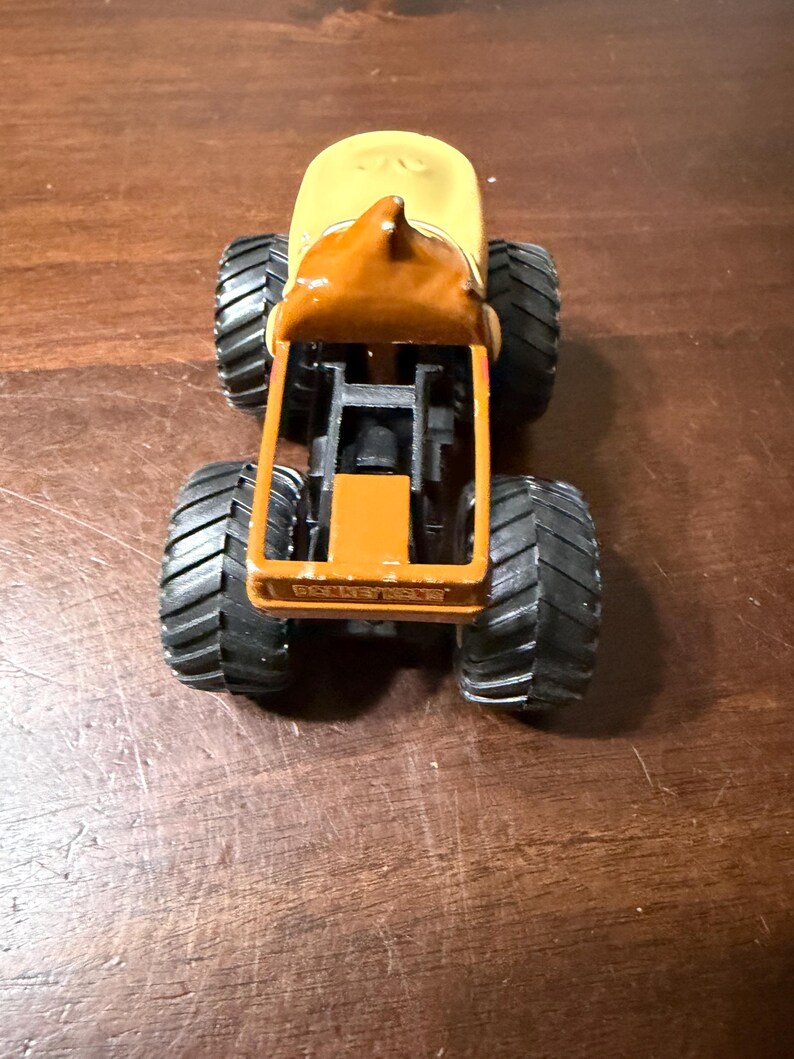 May include: A toy monster truck with a yellow cab and a brown roof. The truck has large black tyres and a brown chassis. The truck is sitting on a wooden surface.