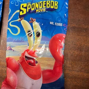 May include: A blue package featuring the SpongeBob Movie. The package displays a cartoon image of Mr. Krabs, with yellow eyes and a red body. The text "ONLY IN THEATRES" and "MR. KRABS" are also visible.