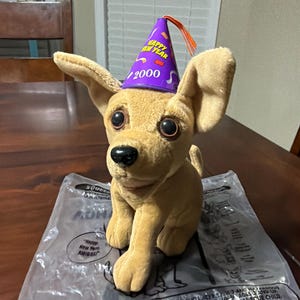 May include: A plush tan chihuahua dog wearing a purple party hat with "Happy New Year 2000" printed on it. The dog is sitting on a plastic bag with text that reads "Happy New Year AMIGOS".