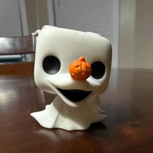 Könnte beinhalten: Eine weiße Vinylfigur eines Charakters aus The Nightmare Before Christmas, mit großen schwarzen Augen und einer orangefarbenen Kürbisnase. Die Figur hat einen breiten, lächelnden Mund und einen fließenden weißen Körper.