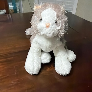 May include: A plush toy cat with a white face and paws, and a gray and white body. The cat has a pink nose and long white whiskers. The toy is sitting on a dark brown surface.