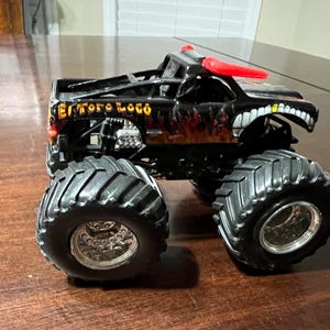 Op de afbeelding: Zwart monstertruckspeelgoed met grote banden en de woorden "El Toro Loco" in gouden letters. De truck heeft een vlammenontwerp en een witte grijns met tanden aan de voorkant. Rode hoorns zijn op het dak gemonteerd.