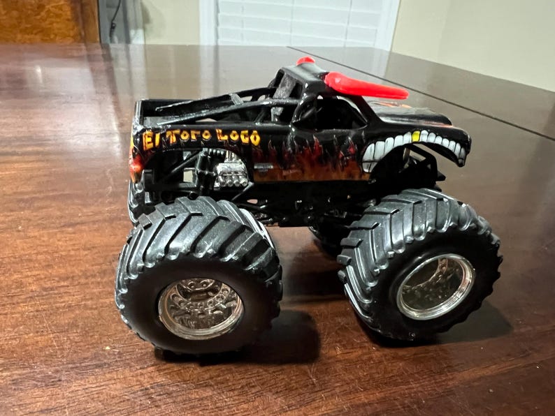 Op de afbeelding: Zwart monstertruckspeelgoed met grote banden en de woorden "El Toro Loco" in gouden letters. De truck heeft een vlammenontwerp en een witte grijns met tanden aan de voorkant. Rode hoorns zijn op het dak gemonteerd.