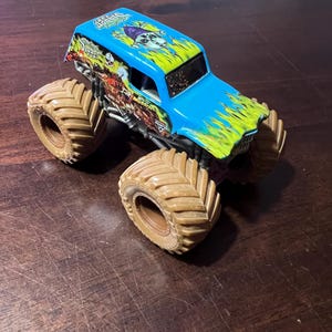 May include: A blue and green toy monster truck with large brown tires. The truck has a skull graphic and the word "Digger" on the roof. The truck is on a wooden surface.