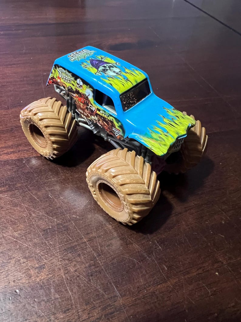 May include: A blue and green toy monster truck with large brown tires. The truck has a skull graphic and the word "Digger" on the roof. The truck is on a wooden surface.