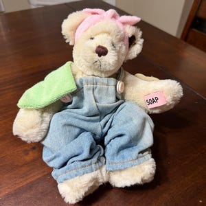May include: A plush teddy bear wearing blue denim overalls, a pink headband, and holding a green washcloth. The bear has a pink tag that reads "SOAP". The bear is sitting on a dark brown wooden surface.