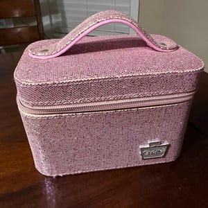 May include: A rectangular pink glittery vanity case with a top handle and a zipper closure. The case features a textured surface and a silver-toned metal emblem on the front with the text "Cookies" embossed in the center of the logo.