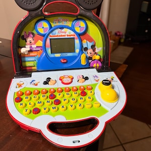 Puede incluir: Un juguete de computadora portátil VTech Disney Mickey Mouse Clubhouse Mousekadoer con un marco negro en forma de Mickey, una pantalla LCD central, un teclado QWERTY colorido con botones de letras y números y un joystick amarillo.