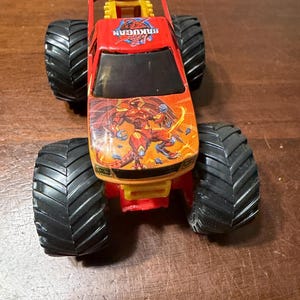 K&ouml;nnte beinhalten: Ein roter und orangefarbener Spielzeug-Monstertruck mit gro&szlig;en schwarzen Reifen. Der Truck tr&auml;gt den Text "Bakugan" auf der Motorhaube und eine Grafik einer Kreatur. Das Spielzeug ist aus Kunststoff und hat ein gelbes Fahrgestell.