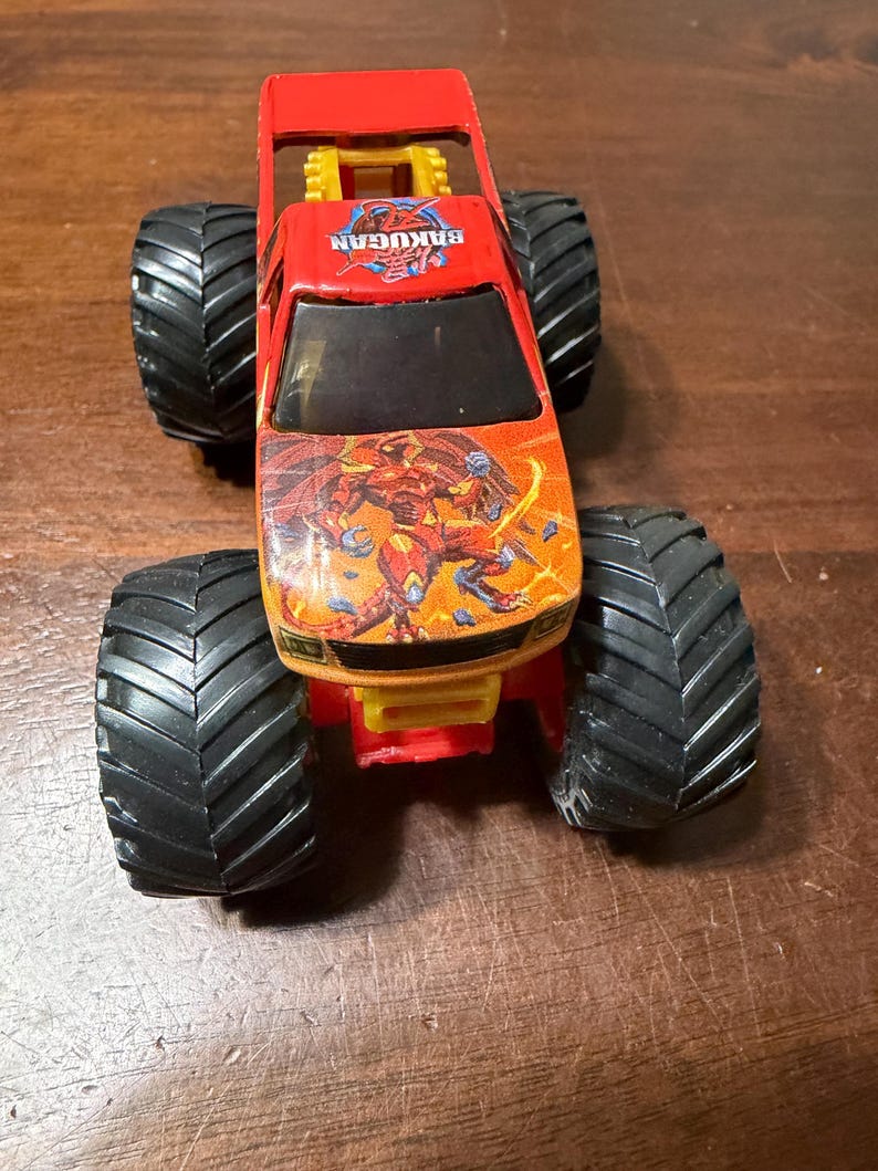 K&ouml;nnte beinhalten: Ein roter und orangefarbener Spielzeug-Monstertruck mit gro&szlig;en schwarzen Reifen. Der Truck tr&auml;gt den Text "Bakugan" auf der Motorhaube und eine Grafik einer Kreatur. Das Spielzeug ist aus Kunststoff und hat ein gelbes Fahrgestell.