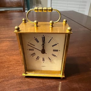 May include: A gold-colored quartz clock with a rectangular shape and a handle. The clock face is cream-colored with black Roman numerals and hands. The clock is sitting on a dark wooden surface.