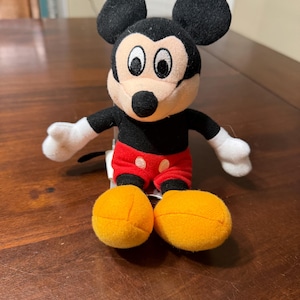 May include: A plush Mickey Mouse toy with black ears, a black body, white gloves, red shorts with white polka dots, and large yellow feet. The toy is sitting on a wooden surface.