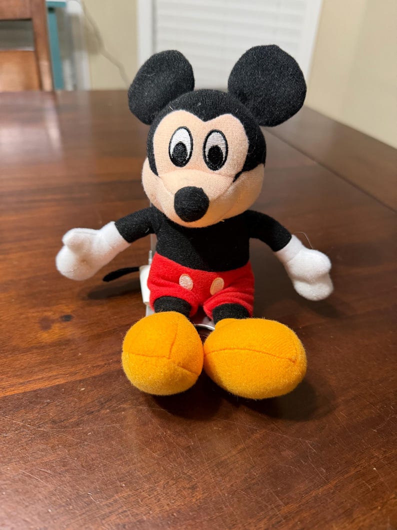 May include: A plush Mickey Mouse toy with black ears, a black body, white gloves, red shorts with white polka dots, and large yellow feet. The toy is sitting on a wooden surface.