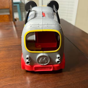 K&ouml;nnte beinhalten: Ein grauer und roter Spielzeugbus mit schwarzen Mickey Mouse Ohren oben. Der Bus hat gelbe und rote Akzente, einen roten Innenraum und das Fisher-Price-Logo. Das Spielzeug steht auf einer braunen Holzoberfl&auml;che.