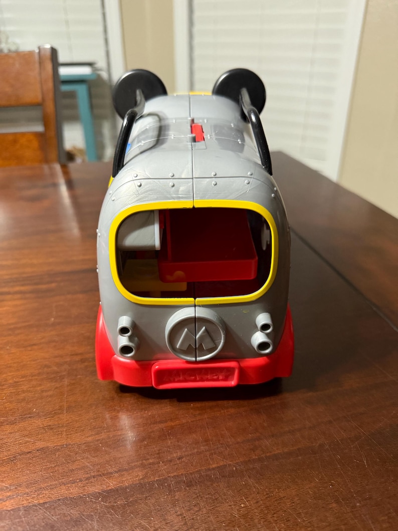 K&ouml;nnte beinhalten: Ein grauer und roter Spielzeugbus mit schwarzen Mickey Mouse Ohren oben. Der Bus hat gelbe und rote Akzente, einen roten Innenraum und das Fisher-Price-Logo. Das Spielzeug steht auf einer braunen Holzoberfl&auml;che.