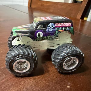 May include: A black and green Grave Digger monster truck toy with large black tires and a glowing chassis. The truck features the Grave Digger logo and a skull graphic. The toy is on a wooden surface.