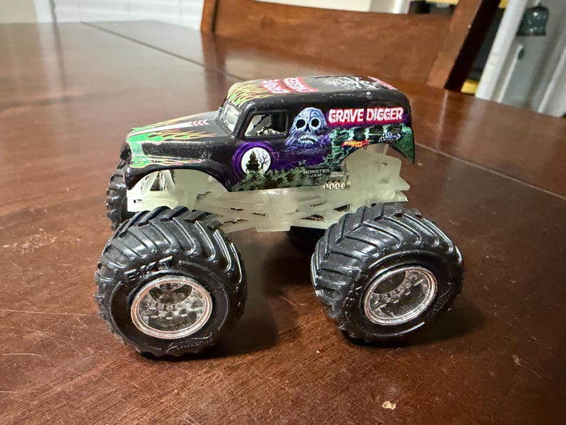 May include: A black and green Grave Digger monster truck toy with large black tires and a glowing chassis. The truck features the Grave Digger logo and a skull graphic. The toy is on a wooden surface.
