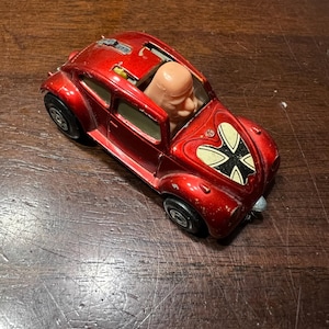 May include: A red toy car with a white cross on the bonnet and a plastic figure of a man in the driver's seat.