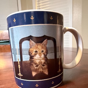 May include: A ceramic coffee mug with a blue rim and handle. The mug features a detailed illustration of a tabby cat sitting at a table, with its reflection visible. The design includes gold accents and a light blue background.