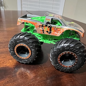 May include: A toy monster truck with large black tires and a green chassis. The truck's body is orange and silver with a weathered design and the text "Hot Wheels" and "Truck" on the side.