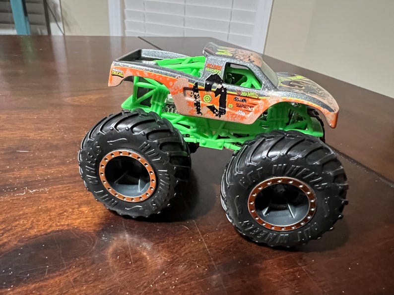 May include: A toy monster truck with large black tires and a green chassis. The truck's body is orange and silver with a weathered design and the text "Hot Wheels" and "Truck" on the side.